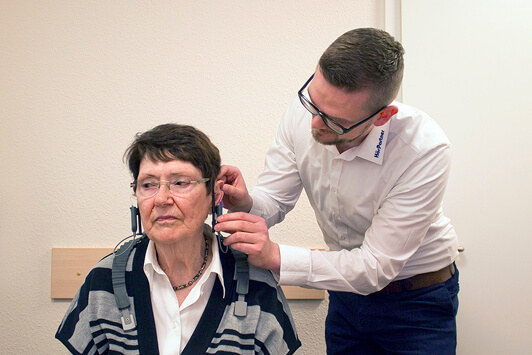 Ein Hörakustiker passt einer älteren Frau ein Hörgerät im Hörtest-Raum an.